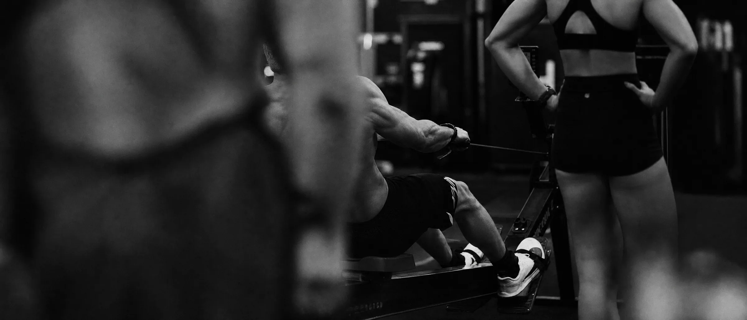 A man on a rowing machine in a gym, a training partner standing beside him